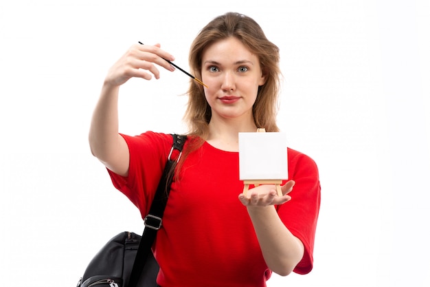 A front view young female student in red shirt black bag holding paintbrush painting smiling on the white
