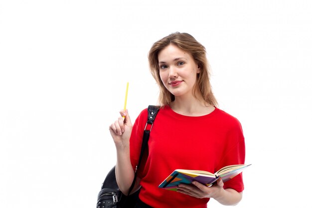Вид спереди молодая студентка в красной рубашке черная сумка, держа перо и тетради, улыбаясь на белом