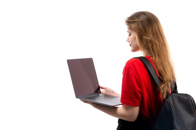Una giovane studentessa di vista frontale nella borsa rossa del nero della camicia facendo uso del computer portatile sul bianco