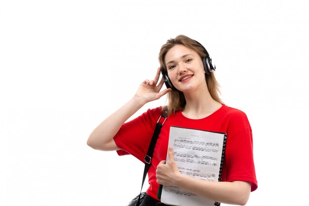 Вид спереди молодая студентка в красной рубашке черная сумка с черными наушниками, слушая музыку, улыбаясь, держа тетрадь на белом