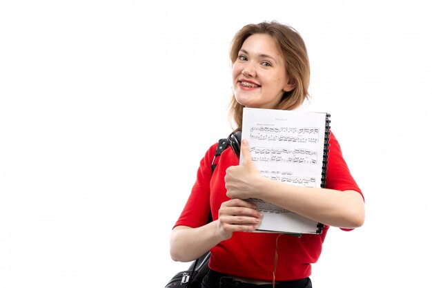 A front view young female student in red shirt black bag with black earphones smiling holding copybook on the white