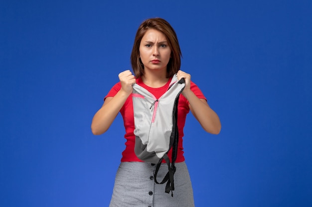 Front view young female student in red shirt holding grey backpack on light-blue background.