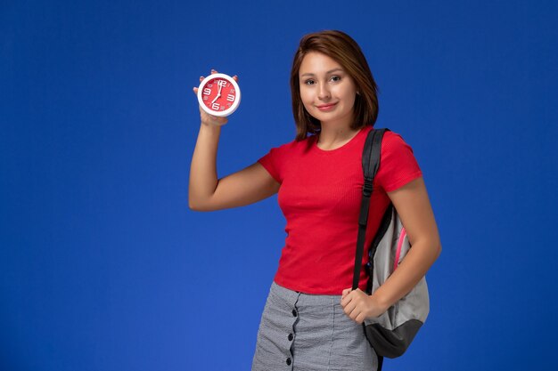 Front view young female student in red shirt wearing backpack holding clocks on light blue background.