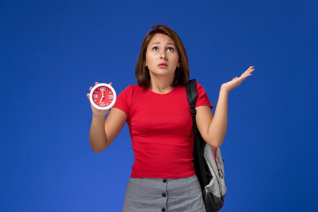 水色の背景に時計を保持しているバックパックを身に着けている赤いシャツの正面図若い女子学生。