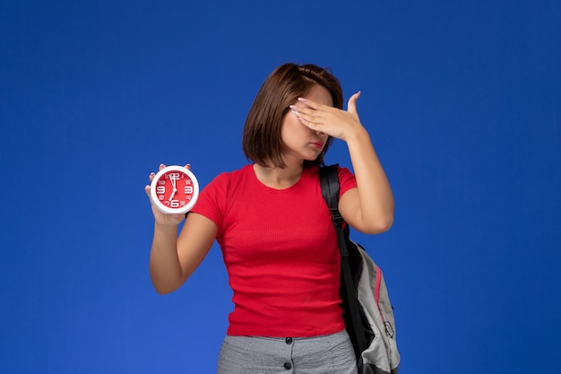 Вид спереди молодая студентка в красной рубашке носить рюкзак, держа часы на светло-синем фоне.