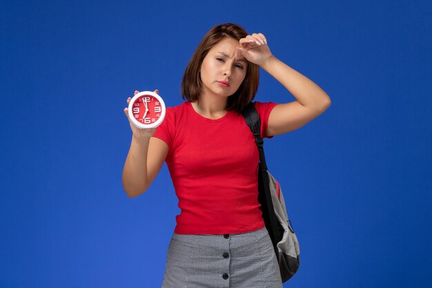 Front view young female student in red shirt wearing backpack holding clocks on the light blue background.