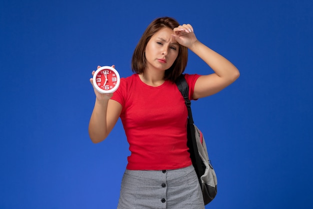 Front view young female student in red shirt wearing backpack holding clocks on the light blue background.