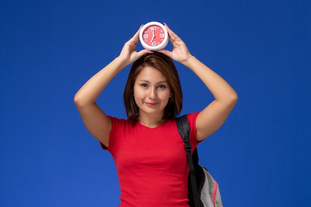 Foto gratuita vista frontale giovane studentessa in camicia rossa che indossa uno zaino che tiene gli orologi sorridente su sfondo azzurro.