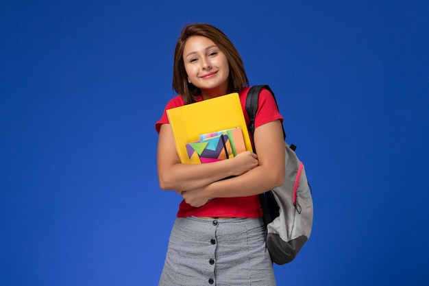 Front view young female student in red shirt wearing backpack holding copybook and files on light blue background.