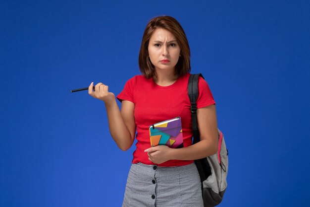 Вид спереди молодая студентка в красной рубашке носить рюкзак, держа тетрадь на светло-синем фоне.