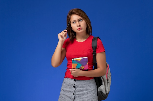 Front view young female student in red shirt wearing backpack holding copybook thinking on the blue background.