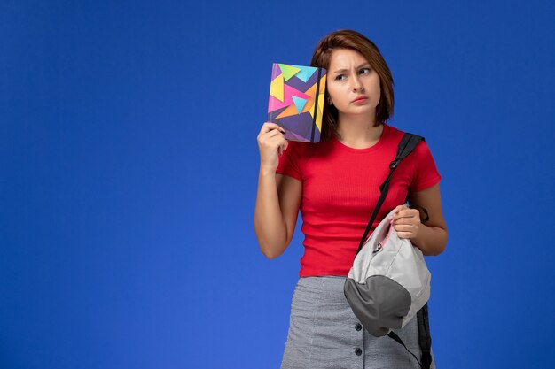 Молодая студентка вид спереди в красной рубашке нося рюкзак, держа тетрадь с прописями на голубом фоне.