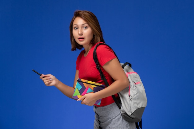 밝은 파란색 배경에 펜으로 카피 북을 들고 배낭을 입고 빨간 셔츠에 전면보기 젊은 여성 학생.