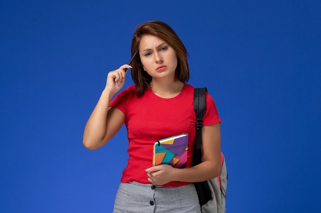 Front view young female student in red shirt wearing backpack holding copybook with pen on light blue background.