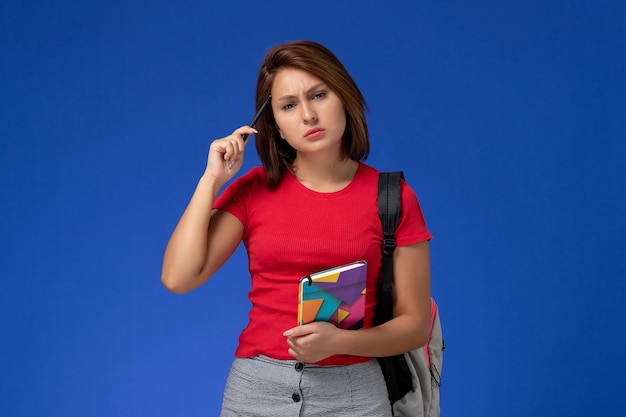 水色の背景にペンでコピーブックを保持しているバックパックを身に着けている赤いシャツの正面図若い女子学生。
