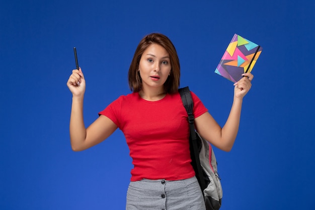 Front view young female student in red shirt wearing backpack holding copybook with pen on light blue desk.