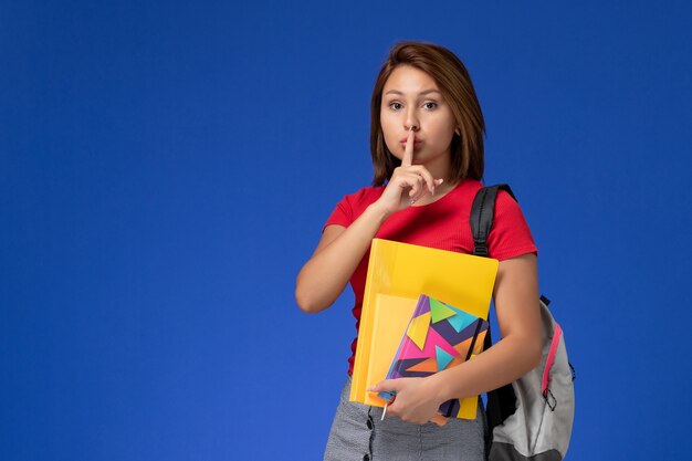 Молодая студентка вид спереди в красной рубашке нося рюкзак, держа файлы и тетрадь на голубом фоне.