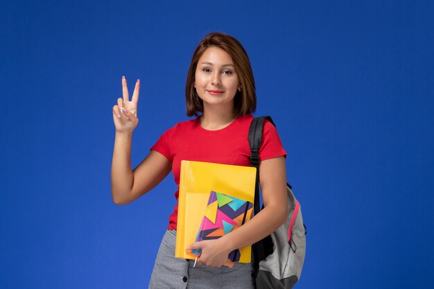 Front view young female student in red shirt wearing backpack holding files and copybook posing on blue background.