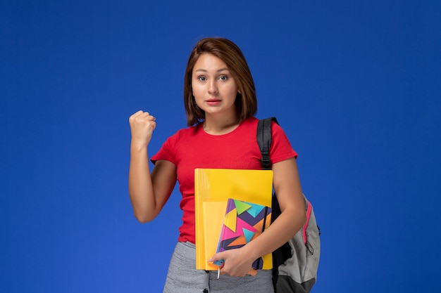ファイルと青い背景で喜んでコピーブックを保持しているバックパックを身に着けている赤いシャツを着た若い女子学生の正面図。