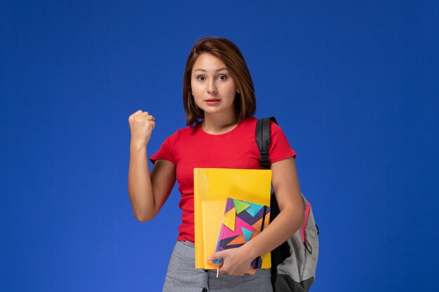 Молодая студентка вид спереди в рюкзаке красной рубашки нося держа файлы и ликование тетради на голубой предпосылке.