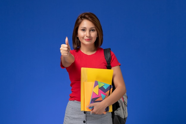 ファイルと青い背景に笑みを浮かべてコピーブックを保持しているバックパックを身に着けている赤いシャツを着た若い女子学生の正面図。