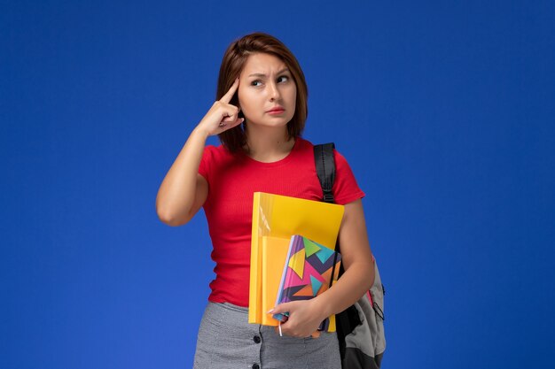 Front view young female student in red shirt wearing backpack holding files and copybook thinking on the blue background.