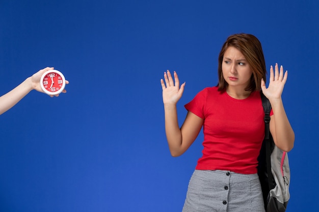 Front view young female student in red shirt wearing backpack posing on light blue background.