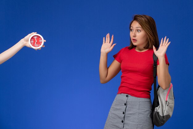 Front view young female student in red shirt wearing backpack scared of clocks on light blue background.