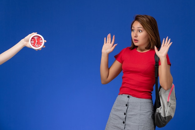 Вид спереди молодая студентка в красной рубашке носить рюкзак испуганные часов на светло-синем фоне.