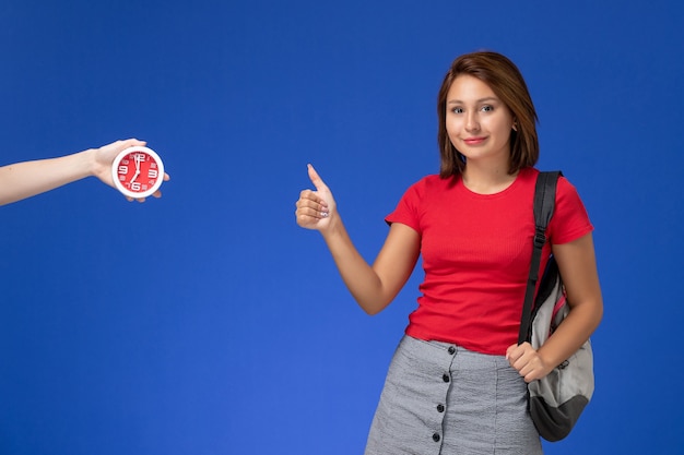 Вид спереди молодая студентка в красной рубашке носить рюкзак, показывая как знак на светло-синем фоне.