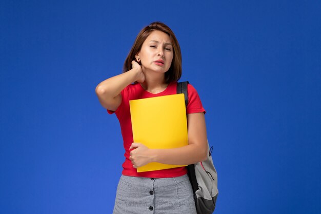 Front view young female student in red shirt with backpack holding yellow files having neckache on light-blue background.