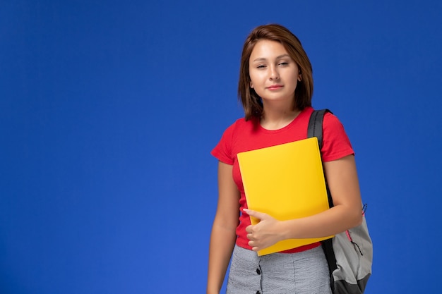 Front view young female student in red shirt with backpack holding yellow files on light blue background.