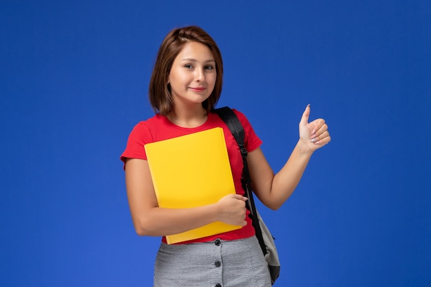 밝은 파란색 배경에 노란색 파일을 들고 배낭 빨간색 셔츠에 전면보기 젊은 여성 학생.