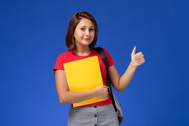 Молодая студентка вид спереди в красной рубашке с рюкзаком, держащим желтые файлы на голубом фоне.