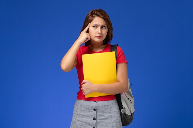 배낭에 노란색 파일을 들고 밝은 파란색 배경에 생각 빨간색 셔츠에 전면보기 젊은 여성 학생.