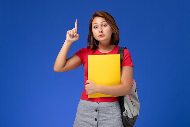 Front view young female student in red shirt with backpack holding yellow files with raised finger on blue background.