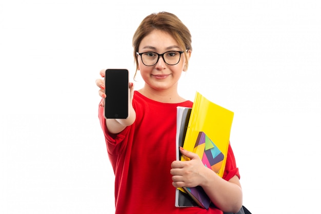 A front view young female student in red t-shirt black jeans holding copybooks showing smartphone on the white