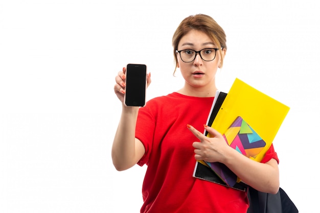 A front view young female student in red t-shirt black jeans holding copybooks smiling showing smartphone on the white