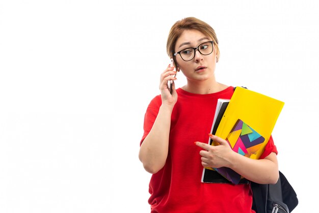 A front view young female student in red t-shirt black jeans holding copybooks talking on the phone on the white