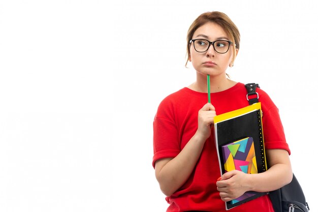 Вид спереди молодая студентка в красной футболке черные джинсы держит различные тетради и файлы, держа карандаш мышления на белом