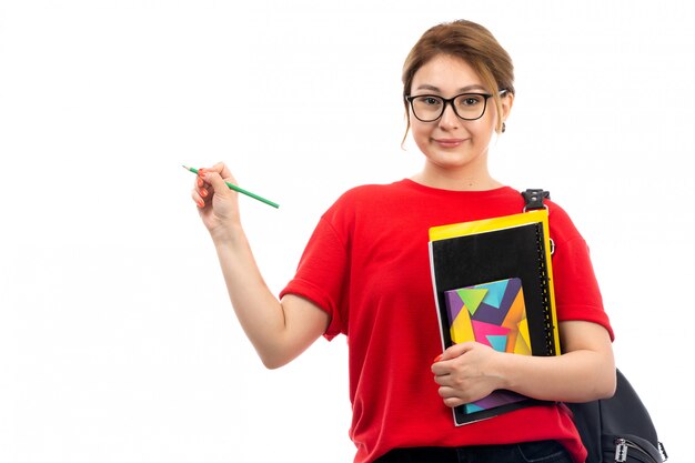 A front view young female student in red t-shirt black jeans holding different copybooks and files holding pencil with bag on the white
