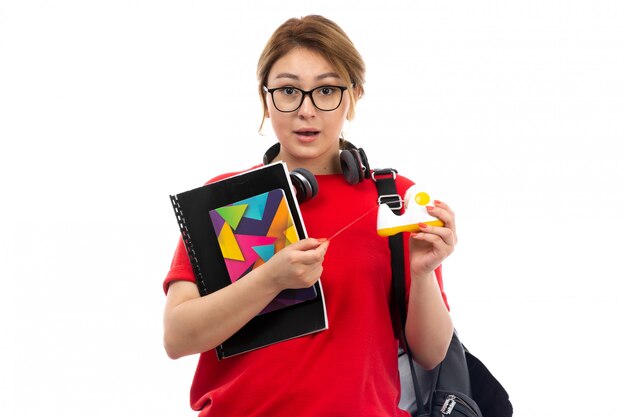 A front view young female student in red t-shirt black jeans wearing black earphones holding copybooks on the white