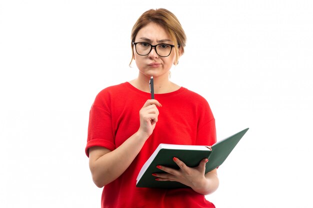 Вид спереди молодая студентка в красной футболке держит тетрадь записывать заметки, думая на белом