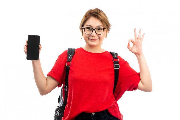 A front view young female student in red t-shirt wearing black bag holding black smartphone smiling on the white