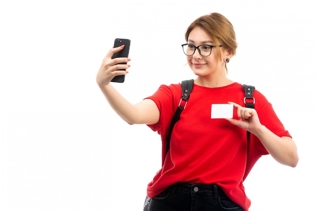白に笑みを浮かべてselfieを取って黒いスマートフォンを保持している黒いバッグを身に着けている赤いtシャツの正面の若い女性学生