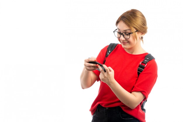 A front view young female student in red t-shirt wearing black bag holding black smartphone using smiling on the white