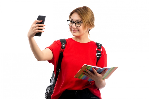 A front view young female student in red t-shirt wearing black bag holding copybook and black smartphone taking a selfie the white