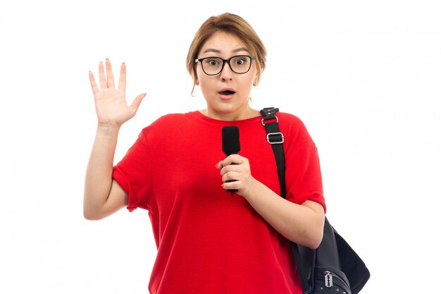 A front view young female student in red t-shirt wearing black bag holding microphone surprised on the white