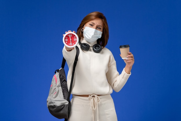 Front view young female student in white jersey wearing mask and backpack holding coffee and clock on blue wall