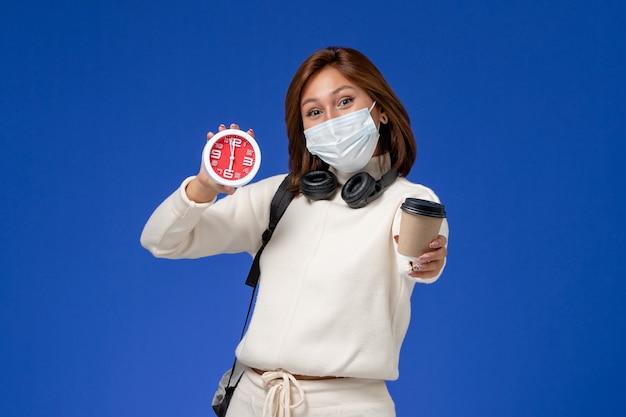 Front view young female student in white jersey wearing mask and backpack holding coffee and clock on the blue wall